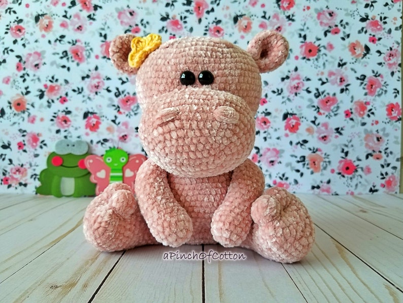 May include: A handmade, plush pink hippo with black eyes and a yellow bow. The hippo is crocheted and sitting upright. The background features a floral pattern and a wooden surface. The text "apinchofcotton" is visible.