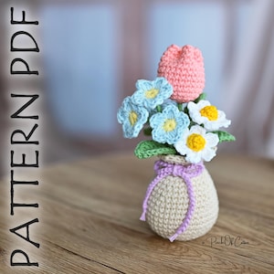 May include: A crocheted bouquet of flowers in a beige vase. The bouquet includes a pink tulip, blue and white flowers with yellow centers, and green leaves. The vase is tied with a lavender ribbon. Text on the left side says "PATTERN PDF".
