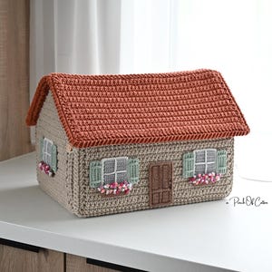 以下が含まれることがあります： テラコッタ色の屋根とベージュの壁が特徴の、かぎ針編みの家型ボックス。家には緑色のシャッター、白いフレームの窓、フラワーボックスがあります。ドアは茶色です。全体的に、手作りの魅力的な装飾品です。