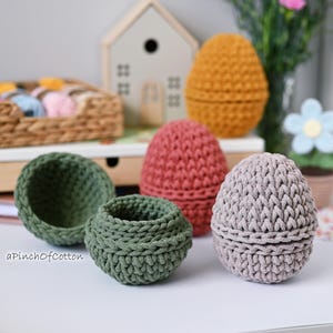 May include: Five crocheted egg-shaped baskets in different colors. The baskets are made of cotton yarn and have a textured surface. The baskets are arranged on a white surface.