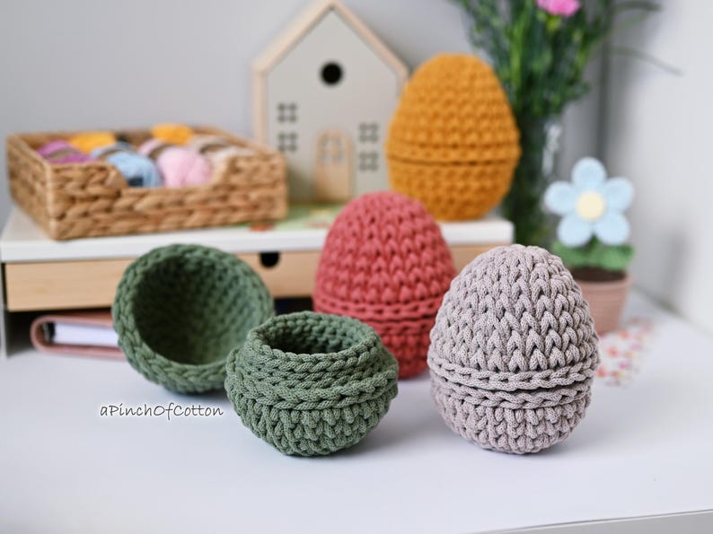 May include: Five crocheted egg-shaped baskets in different colors. The baskets are made of cotton yarn and have a textured surface. The baskets are arranged on a white surface.