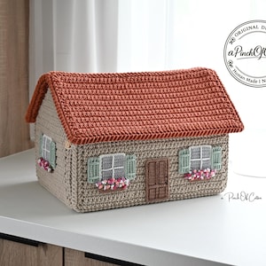 May include: A house-shaped crochet storage box featuring a textured terracotta-colored roof, beige walls, and sage green shutters. The front displays a brown door and windows with pink and white floral accents. Includes a logo reading aPinchOfCotton.