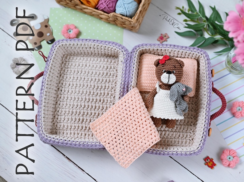 May include: Crocheted suitcase with a teddy bear wearing a white dress and holding a gray bunny. The suitcase is open, with a light pink pillow and blanket inside. The suitcase is lavender and beige. The text "PATTERN PDF" is on the left side.