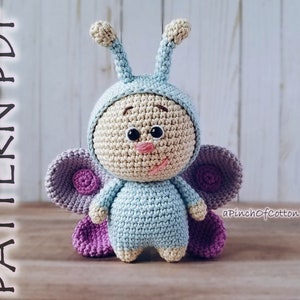May include: A crocheted butterfly toy with blue wings, purple accents, and a smiling face. The toy is wearing a blue and white crocheted costume. The text "aPinchOfCotton" is visible in the bottom right corner of the image.