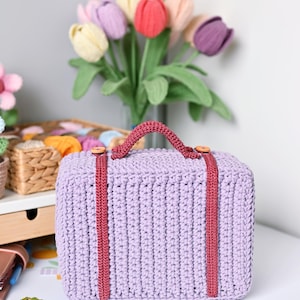 May include: A lavender crochet suitcase with a burgundy handle and straps. The suitcase has wooden button accents and is sitting on a white surface. A bouquet of tulips in various colors is in the background.
