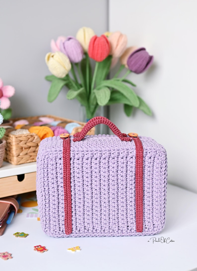 May include: A lavender crochet suitcase with a burgundy handle and straps. The suitcase has wooden button accents and is sitting on a white surface. A bouquet of tulips in various colors is in the background.