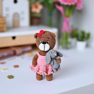 May include: A handmade crochet bear doll with a pink dress and a red bow. The bear is holding a small gray crochet bunny. The bear is brown with a white muzzle and black eyes. A charming, handcrafted toy.
