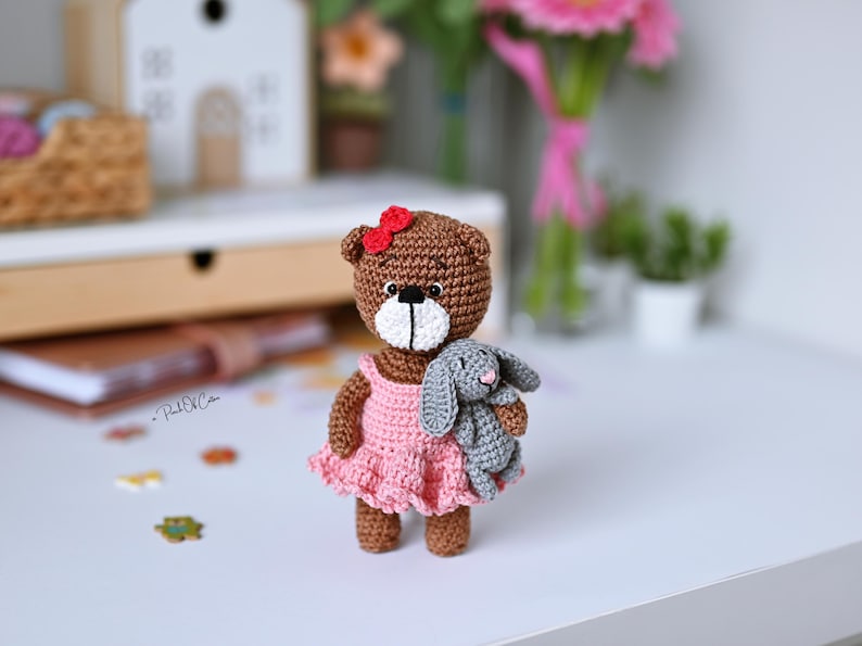 May include: A handmade crochet bear doll with a pink dress and a red bow. The bear is holding a small gray crochet bunny. The bear is brown with a white muzzle and black eyes. A charming, handcrafted toy.
