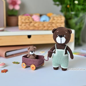 K&ouml;nnte beinhalten: Ein brauner geh&auml;kelter Teddyb&auml;r, der eine hellgr&uuml;ne geh&auml;kelte Latzhose tr&auml;gt, zieht einen braunen Wagen mit einem kleineren braunen geh&auml;kelten Teddyb&auml;ren darin. Der Wagen hat Holzr&auml;der mit bunten Details.