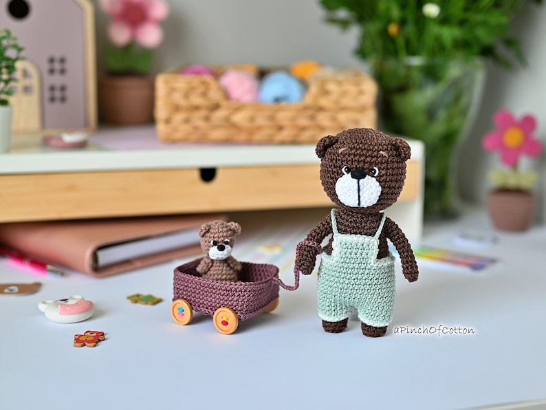 K&ouml;nnte beinhalten: Ein brauner geh&auml;kelter Teddyb&auml;r, der eine hellgr&uuml;ne geh&auml;kelte Latzhose tr&auml;gt, zieht einen braunen Wagen mit einem kleineren braunen geh&auml;kelten Teddyb&auml;ren darin. Der Wagen hat Holzr&auml;der mit bunten Details.