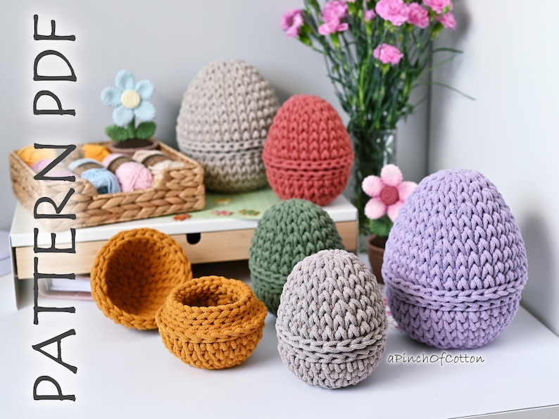 May include: A collection of crocheted egg-shaped baskets in various colors, including purple, gray, green, red, and beige. The baskets are arranged on a white surface with a wooden shelf in the background. The text "aPinchOfCotton" is visible in the lower right corner of the image.