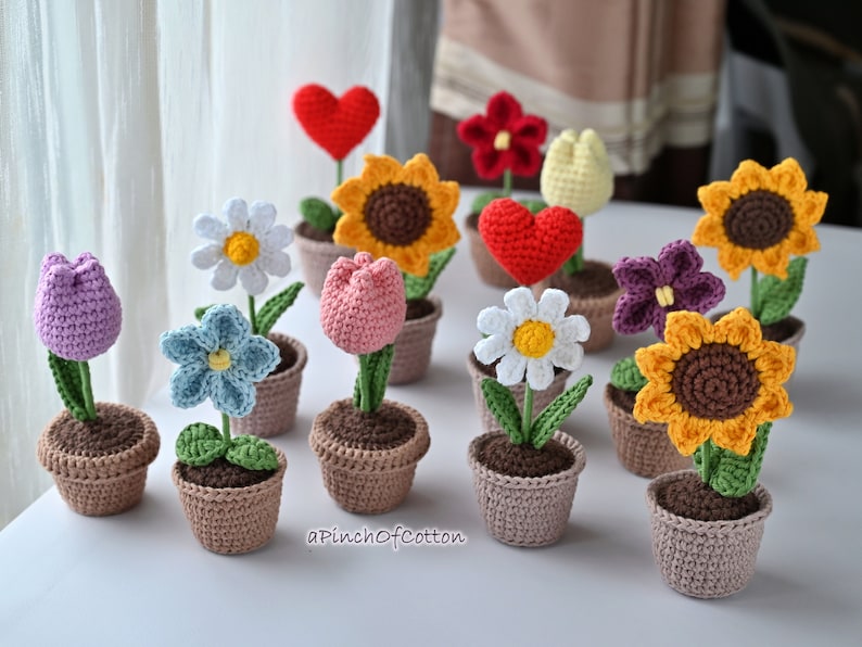 May include: A collection of 11 crocheted flower arrangements in various colors, including purple, pink, blue, white, red, and yellow. Each flower is potted in a brown crocheted pot. The flowers are arranged on a white surface with the text "a Pinch of Cotton" visible in the bottom right corner.