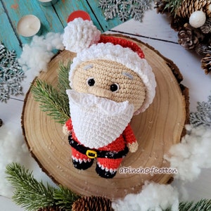 May include: A crocheted Santa Claus doll with a white beard and red and white hat. The doll is sitting on a wooden slice with a white background.