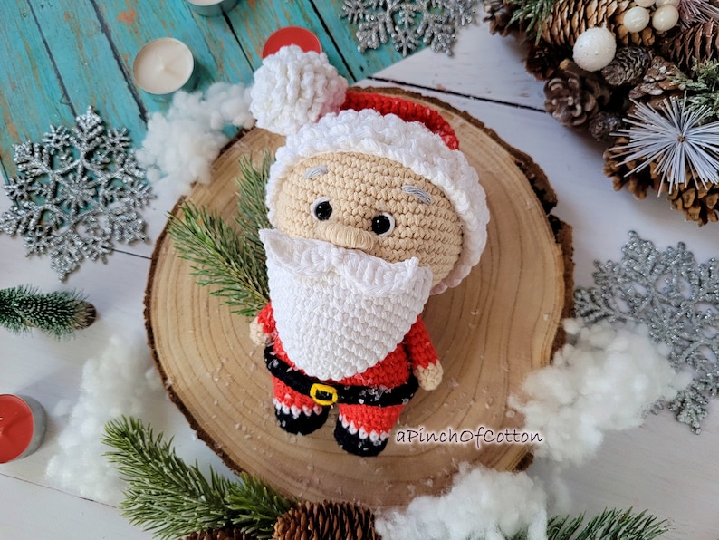 May include: A crocheted Santa Claus doll with a white beard and red and white hat. The doll is sitting on a wooden slice with a white background.