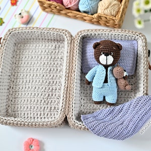 K&ouml;nnte beinhalten: Ein geh&auml;kelter Koffer mit einem braunen Teddyb&auml;r, der einen blauen Schlafanzug tr&auml;gt. Der Koffer ist ge&ouml;ffnet und der Teddyb&auml;r liegt auf einer lila Decke. Der Koffer hat einen braunen Griff und ein wei&szlig;es H&auml;kelfutter.