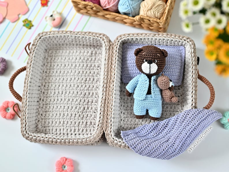 K&ouml;nnte beinhalten: Ein geh&auml;kelter Koffer mit einem braunen Teddyb&auml;r, der einen blauen Schlafanzug tr&auml;gt. Der Koffer ist ge&ouml;ffnet und der Teddyb&auml;r liegt auf einer lila Decke. Der Koffer hat einen braunen Griff und ein wei&szlig;es H&auml;kelfutter.