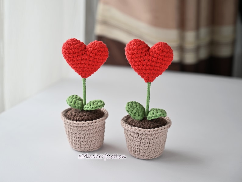 May include: Two crocheted heart-shaped flower pots with green leaves. The pots are beige and the hearts are red.