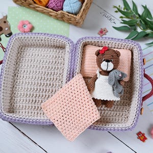 May include: A handmade crochet suitcase in shades of purple, beige, and red, open to reveal a crocheted bed with a teddy bear and a gray bunny. A pink blanket rests beside the bed. The scene is decorated with small crochet flowers and a basket of yarn.