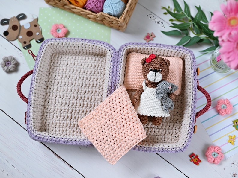 May include: A handmade crochet suitcase in shades of purple, beige, and red, open to reveal a crocheted bed with a teddy bear and a gray bunny. A pink blanket rests beside the bed. The scene is decorated with small crochet flowers and a basket of yarn.