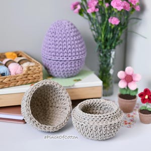 May include: Three crocheted baskets in different sizes, one purple and two beige, with a small crocheted flower pot with a red flower and a larger crocheted flower pot with a pink flower. The baskets are on a table with a book and a wicker basket filled with yarn.