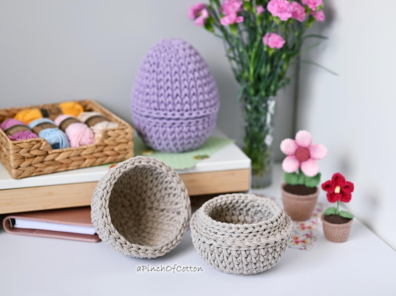 May include: Three crocheted baskets in different sizes, one purple and two beige, with a small crocheted flower pot with a red flower and a larger crocheted flower pot with a pink flower. The baskets are on a table with a book and a wicker basket filled with yarn.
