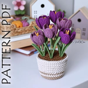 May include: A crocheted purple crocus flower arrangement in a white and brown pot. The flowers have yellow centers and green stems. The pot is sitting on a white surface. The text "PATTERN PDF" is on the left side of the image.