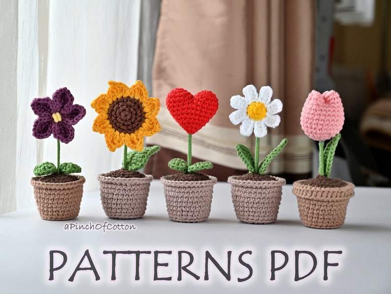 May include: Five crocheted flower arrangements in pots. The flowers are a purple flower, a yellow sunflower, a red heart, a white daisy, and a pink tulip. The pots are all brown.