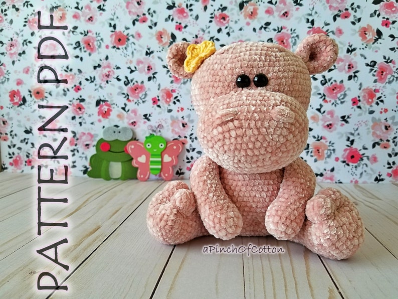 May include: A handmade, plush pink hippo with black eyes and a yellow bow. The hippo is crocheted and sitting down. The background features a floral pattern and two wooden cutouts of a frog and butterfly. The text "PATTERN PDF" is on the left side.