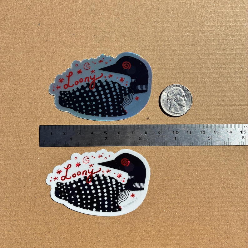 May include: Two die-cut stickers shaped like a loon bird with black and white spotted patterns, red text reading Loony, and red star and moon accents. A metal ruler shows the stickers are approximately 3.5 inches wide, next to a US quarter coin.