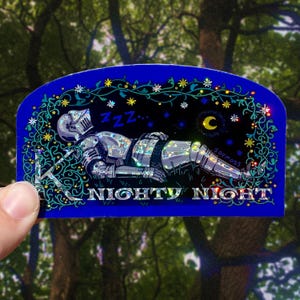 May include: A blue sticker with a sleeping knight in silver armor, surrounded by stars, a crescent moon, and floral accents. The text "NIGHTY NIGHT" is at the bottom. The sticker is held by a hand.
