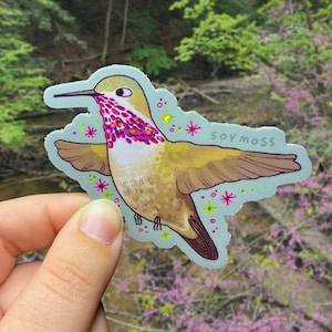 May include: A sticker featuring a hummingbird with outstretched wings. The bird has a yellow body, brown wings, and a red and purple throat. The sticker has a light blue border and the text "soy moss".