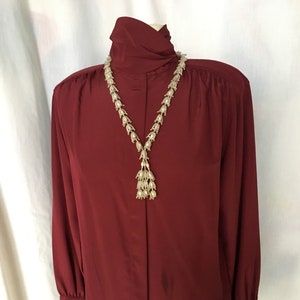 May include: A burgundy long-sleeved blouse with a high neck and a beaded necklace with a tassel.