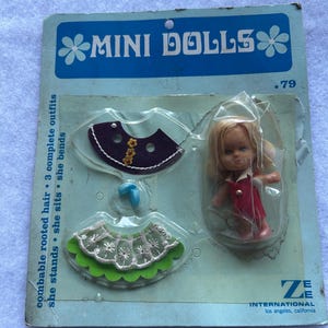 May include: A vintage Mini Dolls blister pack featuring a small doll with blonde hair wearing a red dress, accompanied by two additional outfits: a purple skirt with yellow floral accents and a green skirt with white lace trim, plus a blue accessory.