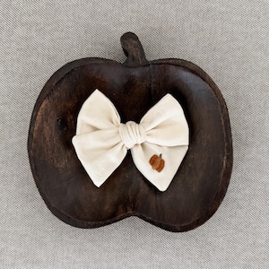 May include: A cream-coloured bow with a small embroidered pumpkin detail, resting on a dark brown, pumpkin-shaped wooden dish. The bow is centred, and the dish is on a grey textured surface.