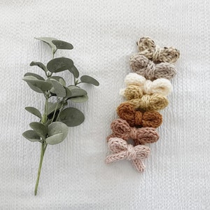May include: Set of eight knitted hair bows in various shades of brown, beige, and cream. The bows are arranged in a vertical stack, with the darkest shade at the bottom and the lightest shade at the top.