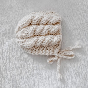 May include: A white knitted baby bonnet with a cable knit pattern and a tie at the back.