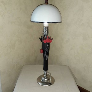 May include: A table lamp made from a Budweiser Select beer tap handle. The lamp has a white lampshade with a black and white checkered trim. The base of the lamp is silver and the tap handle is black with the Budweiser Select logo.