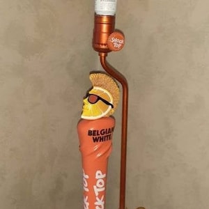 May include: A Shock Top Belgian White beer tap handle lamp with a white lampshade and a lemon slice on top. The tap handle is orange and yellow with the words "Shock Top" and "Belgian White" on it.