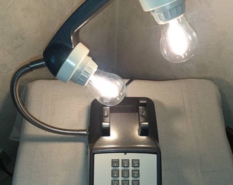 May include: A black rotary phone shaped desk lamp with a flexible arm and two light bulbs. The phone has a keypad with numbers and letters, and a display that reads "067-5309".
