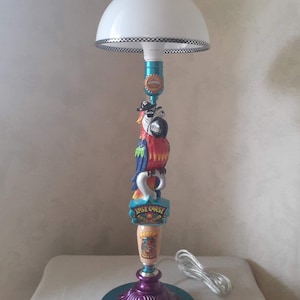 May include: A turquoise and purple table lamp with a white lampshade. The lamp features a parrot figurine with a pirate hat and a "Lost Coast" sign. The lamp is on a white surface.