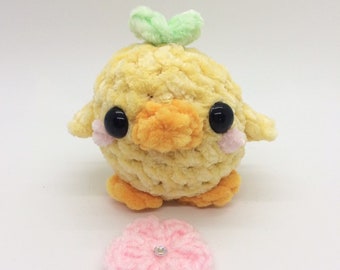 May include: A crocheted yellow duck plush toy with black eyes and a green leaf on its head. The duck is sitting on a white surface with a pink crocheted flower.