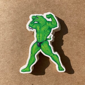 May include: Green, cartoon-style sticker of a muscular, anthropomorphic creature with a long snout, wearing a patterned swimsuit. The creature is flexing its biceps and has a determined expression.