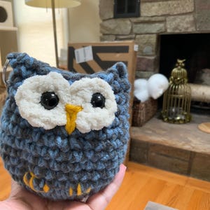 May include: A handmade crochet owl keychain in shades of blue and gray, with white and yellow accents. The owl has large black eyes and a fluffy white chest. A small metal ring is attached to the top.