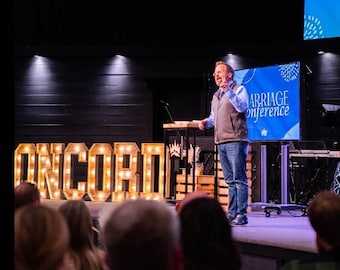 Op de afbeelding: Een man in een blauw vest en jeans spreekt op een podium met een groot verlicht bord met de tekst "CONCORD" ervoor. Het bord is gemaakt van houten letters met gloeilampen. Achter hem is een scherm met de tekst "Marriage Conference".