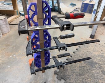 Op de afbeelding: Drie paarse 3D-geprinte kunststof klemblokken zijn verticaal tegen een houten plank geplaatst. Drie metalen lijmklemmen met zwarte en rode handvatten zijn horizontaal over de blokken aangedraaid om de houten delen stevig vast te zetten.