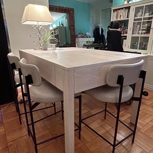 May include: A white wooden kitchen island with a white countertop and black metal bar stools. The island has a drawer and a shelf. The stools have a white padded seat and black metal legs.