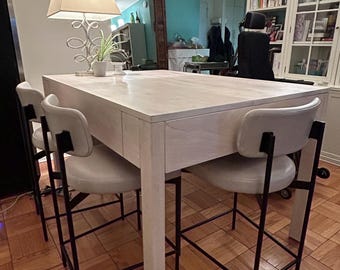 Op de afbeelding: Een wit houten keukeneiland met een wit werkblad en zwarte metalen barstoelen. Het eiland heeft een lade en een plank. De krukken hebben een witte gepolsterde zitting en zwarte metalen poten.