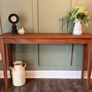 May include: A rectangular wooden console table with four tapered legs. On the surface sits a black round clock, a small white bird figurine, two thin brass candlesticks, and a white metal pitcher filled with green flowers and a tan ribbon.