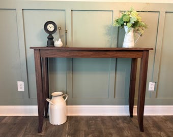 Op de afbeelding: Een rechthoekige donkerbruine houten sidetable met vier taps toelopende poten. Op de tafel staan een zwarte ronde klok, een klein wit keramieken vogeltje en een witte metalen kan gevuld met groene bloemen. Een grote witte metalen melkbus staat eronder.