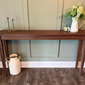 May include: A long, rectangular wooden console table with four tapered legs. On the table surface are two thin brass candlesticks, a small white pumpkin, and a white pitcher filled with green foliage. A large white metal milk can sits below.